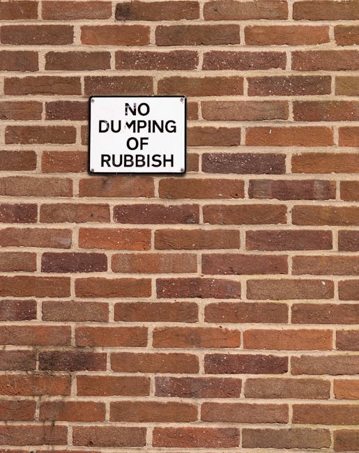 A square white sign with black text reading 'NO DUMPING OF RUBBISH' is mounted on a red brick wall. The wall consists of horizontally laid bricks with a rough texture, in varying shades of red, brown, and orange, separated by light grey mortar joints. The sign is positioned in the upper left quadrant of the image, and the brick surface extends across the entire background, suggesting an outdoor setting. The lighting appears natural, with shadows cast on the lower left side of the wall, highlighting the textured surface of the bricks. This scene illustrates a typical area where private waste removal services might operate, emphasizing the importance of adhering to local rules against illegal dumping and supporting responsible rubbish disposal through professional rubbish removal companies such as Rubbish Removal Bromley.