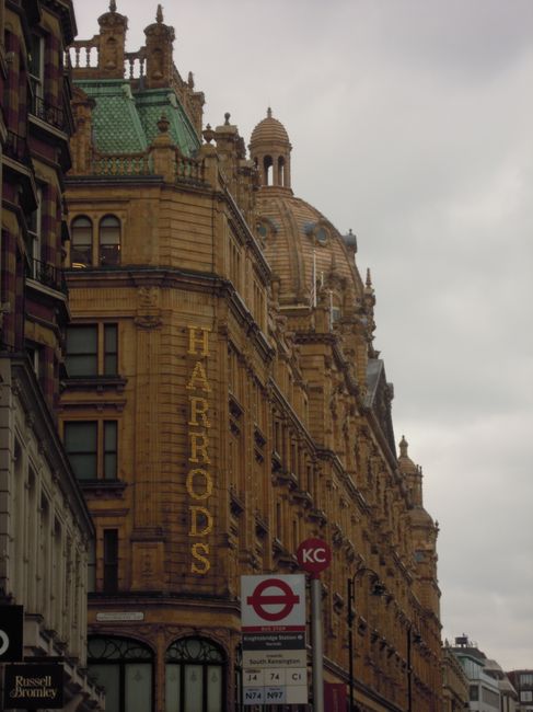 A detailed photograph of a historic Victorian-style building constructed from yellowish-brown stone, featuring ornate architectural details such as decorative cornices, balustrades, and domed towers with finials. The building's façade displays multiple windows with dark frames and a vertically oriented, illuminated sign reading 'HARRORDS' in yellow letters on the left side. In the foreground, there is a red and white underground station sign with a circular logo and a timetable board showing various bus routes. The sky above is overcast with thick gray clouds, creating diffuse lighting that highlights the intricate textures and finishes of the building's stonework, situated in an urban environment next to other modern and historic structures. The scene reflects elements of city life, including public transportation signage, suggesting proximity to a busy commercial or transport hub in central London, suitable for discussing private waste disposal or alternative rubbish removal services like those offered by Rubbish Removal Bromley.