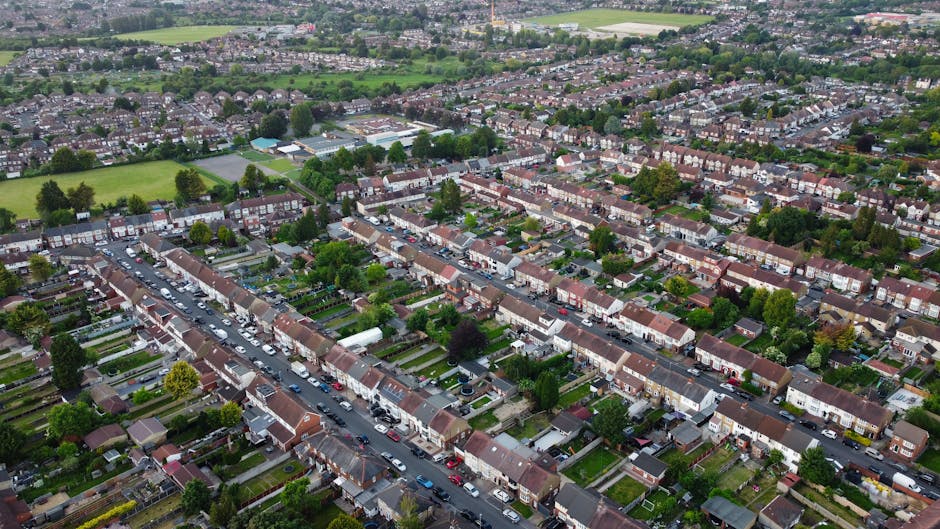 An aerial view of a residential suburb showing numerous rows of terraced houses with tiled roofs and small front gardens, situated along a main street filled with parked cars on both sides. The homes are predominantly brick-built with a variety of colors, including red and brown, with some houses featuring white or grey facades. Lush green trees are interspersed throughout the neighborhood, and several open green spaces, sports fields, and school grounds are visible in the background. The environment is well-maintained, with neatly arranged gardens and paved pathways. The sky is overcast, casting diffuse light over the scene, emphasizing the orderly arrangement of the houses and the surrounding greenery. This suburban setting exemplifies typical private waste collection and rubbish removal contexts, with potential for services like on-site clearance or independent waste disposal by companies such as Rubbish Removal Bromley, catering to household rubbish and estate clearance needs.