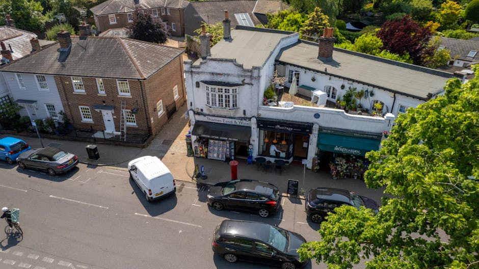 An aerial view of a small commercial street showcasing three retail shops with distinct facades, including a white building with a curved front and decorative architectural details, a shop with a black awning, and another with a green awning. In front of the shops, there are several parked vehicles, including a white van, a black hatchback, and two black cars, positioned along a narrow street with marked parking bays. Nearby, a bicycle is locked to a bicycle rack on the sidewalk, which also features a black trash bin and a red post box. Surrounding the street are residential houses with brick and white exteriors, pitched roofs, and chimneys, set amidst lush green trees providing a vibrant backdrop. The environment suggests a typical small-town setting with mixed-use buildings, and the scene hints at the importance of on-site rubbish management for local businesses, as Rubbish Removal Bromley would facilitate private waste handling for such commercial premises and nearby residences.