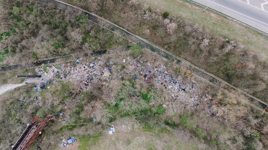 An aerial view of a large, unmanaged area of scattered rubbish and debris, including plastic bags, bottles, cardboard, and other waste materials, spread across a patch of bare earth and sparse vegetation. The rubbish extends from the bottom left towards the center of the image, with some materials piled loosely and others dispersed randomly. To the top right, a paved road runs parallel to a fence that separates the waste area from a strip of grass and trees, which appear to be partly leafless with some green patches. The surrounding environment suggests an outdoor, possibly neglected or abandoned space, with natural light illuminating the scene evenly. This scene visually reflects the impact of illegal dumping or unregulated waste disposal, highlighting the importance of private rubbish removal services like Rubbish Removal Bromley to help manage and clear such sites effectively, maintaining environmental standards and aesthetic appeal in urban or suburban settings.