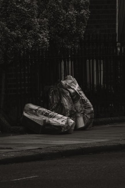 Three large black garbage bags filled with waste material are placed on the pavement beside a black metal fence, which is partially covered by dense foliage. The bags appear to be made of plastic and are crinkled and slightly reflective, indicating they are filled with refuse. The scene is set outdoors on a city street, with the bags positioned near the curb, suggesting an on-site rubbish removal or private waste collection. The background includes a dark, opaque fence and trees with leafy branches, casting shadows over part of the scene. The lighting is subdued, creating a somewhat somber atmosphere typical of early morning or late afternoon urban settings. This image illustrates an example of waste left for collection, possibly managed by a service such as Rubbish Removal Bromley, emphasizing the importance of proper rubbish disposal methods in urban areas, especially when municipal collection is not immediately available.